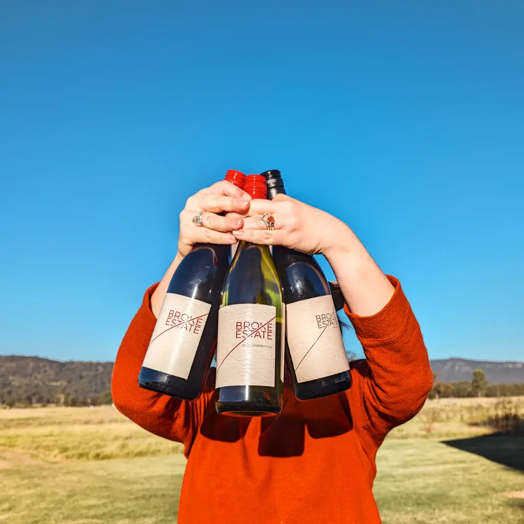 Three bottles of Broke Estate wine held up against a blue sky in a paddock
